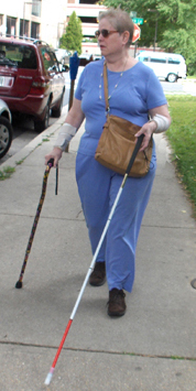 The woman continues to walk on the sidewalk, but this time her left foot is forward, and the tip of the long cane is far in front of her right foot.  The support cane remains close to her right side.